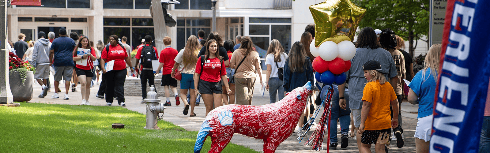 Students walk alongside the sidewalk outside the Campus Center and Perry Buildings on UWG's campus. The Wolf Statue has balloons tied atop it.