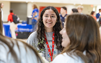 Student smiling at tabling fair while speaking with other students, faculty, and staff.