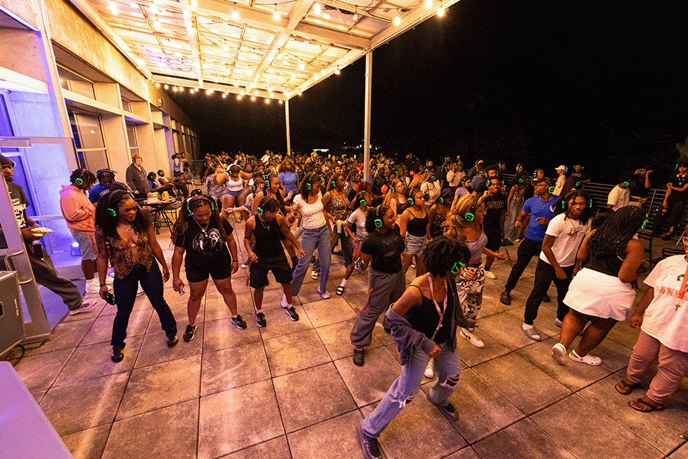 Back to School Balcony Silent Disco: Large group of people wearing headphones dancing together on an outdoor patio under string lights at night.