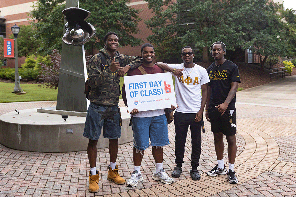 Four people standing on a campus walkway holding a sign that reads "First Day of Class" near a sculpture and campus buildings