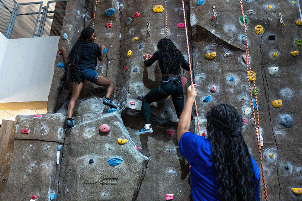 Two people climbing an indoor rock wall with colorful holds while another person stands below holding a rope for safety