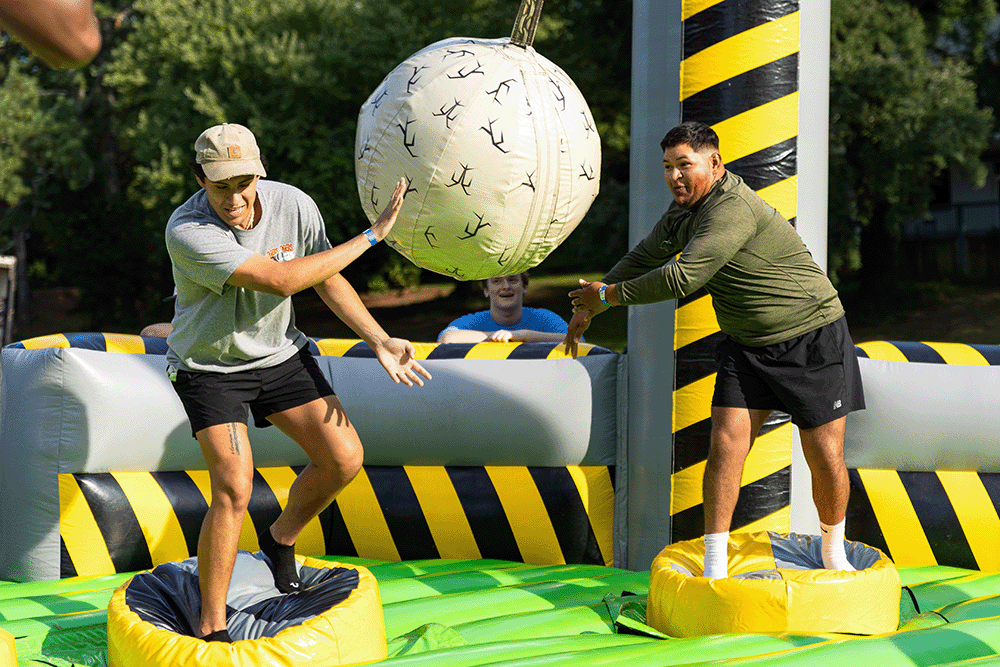 Two people balancing on separate platforms while batting a suspended inflatable ball toward each other on an outdoor inflatable game