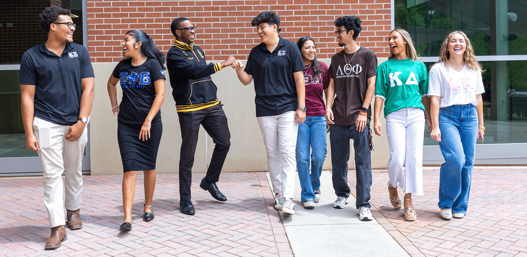 Group of eight diverse college students in Greek-letter shirts from all four councilswalking and talking on a campus path in front of a brick building.