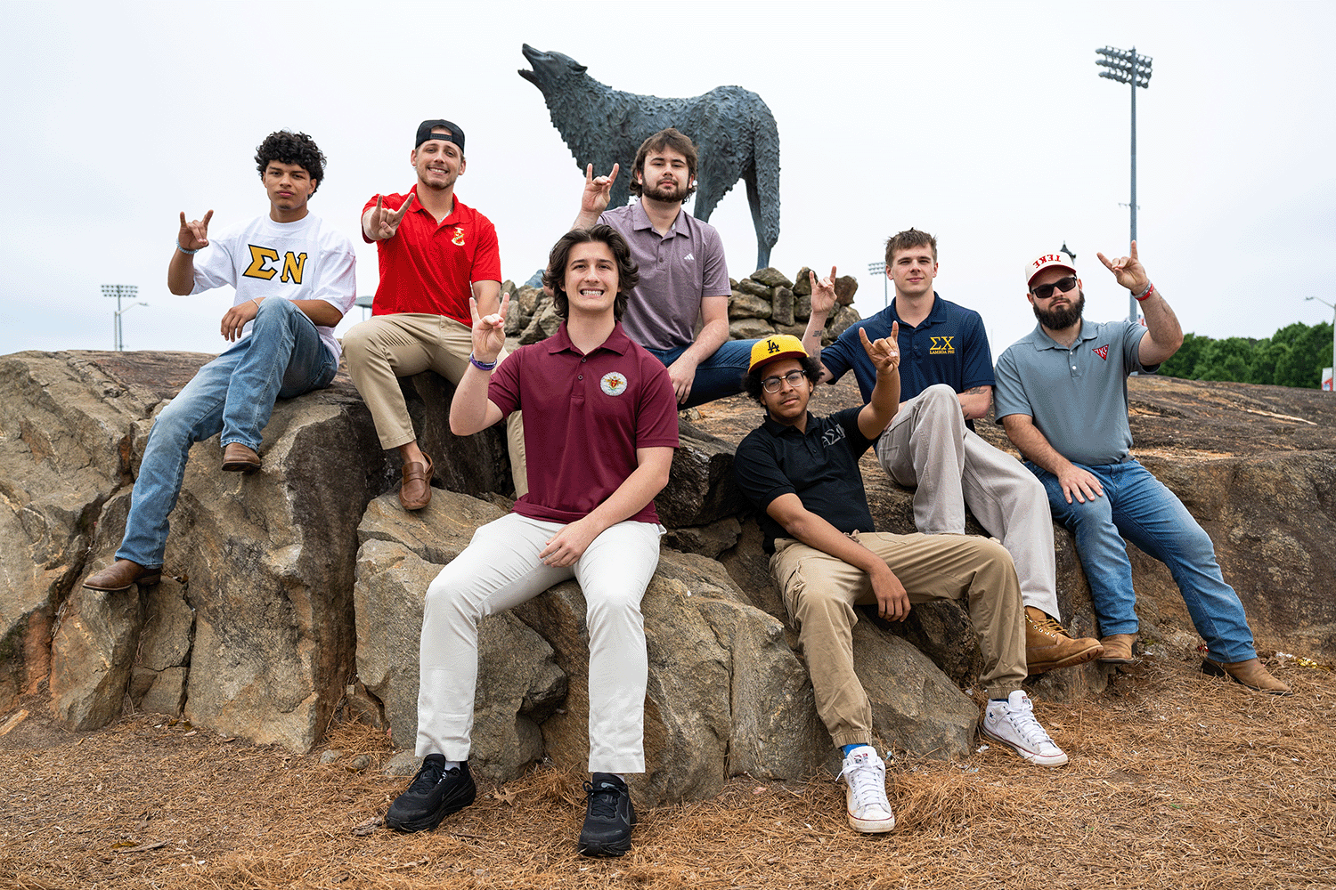 Group of interfraternity council members at wolf statue sitting on rocks holding up wolf signs