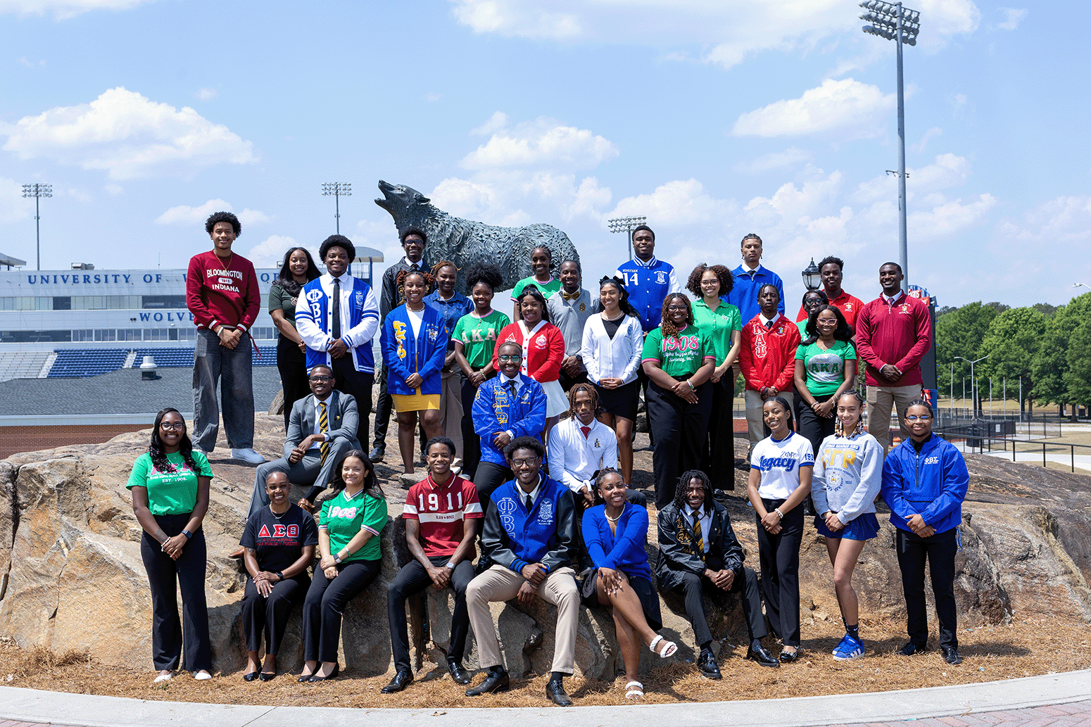 Group of NPHC students posing on rocks in front of a stadium and wolf statue, wearing Greek-letter apparel and smiling at the camera.