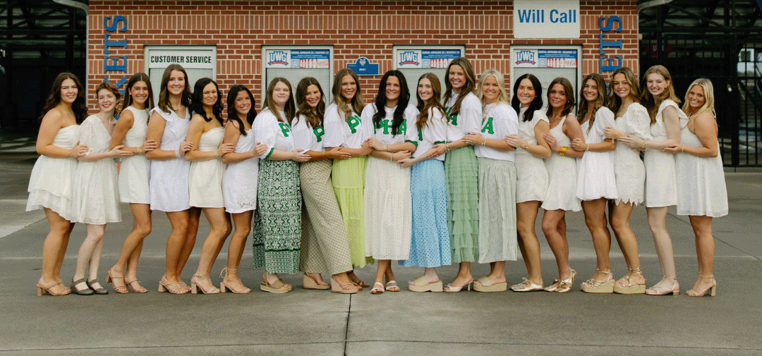 Group of eighteen PHA women standing arm‑in‑arm in front of a brick building, all smiling and dressed in coordinated white and pastel outfits.
