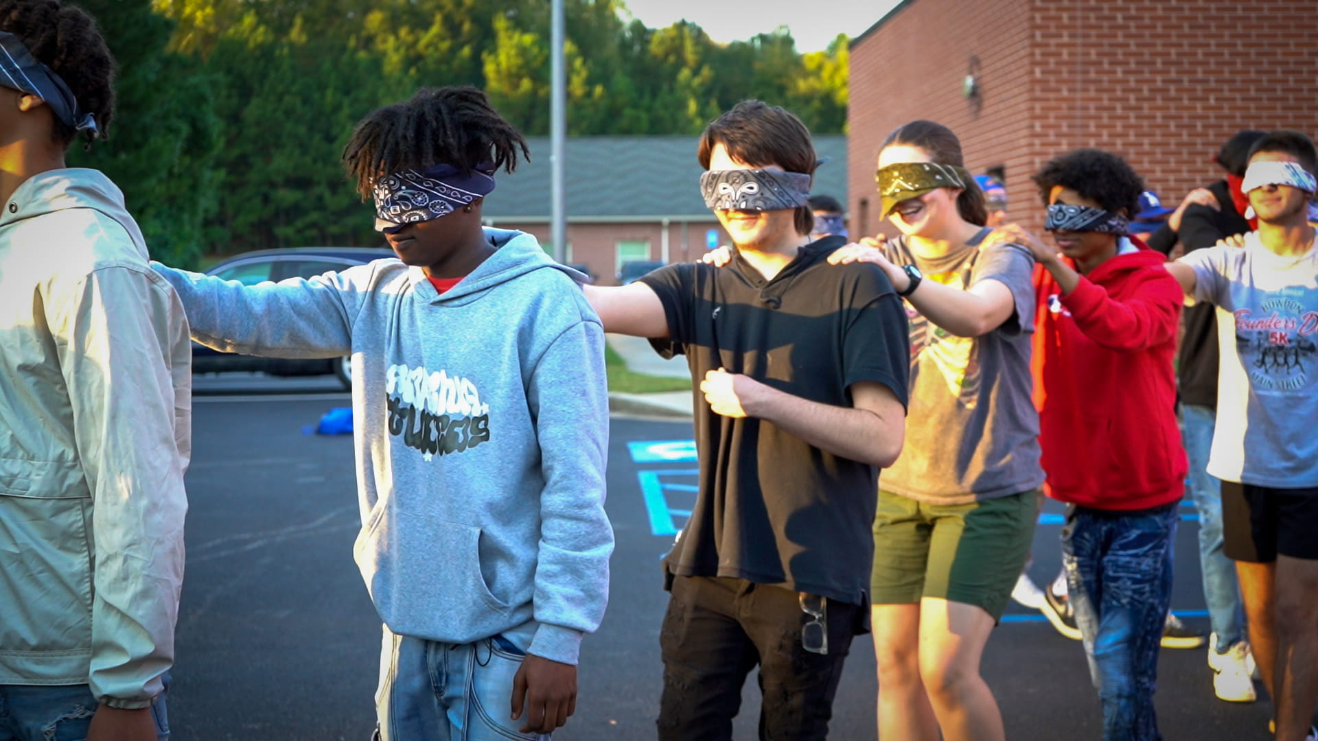 Students blindfolded in a line with their hands on the person's shoulder in front of them.