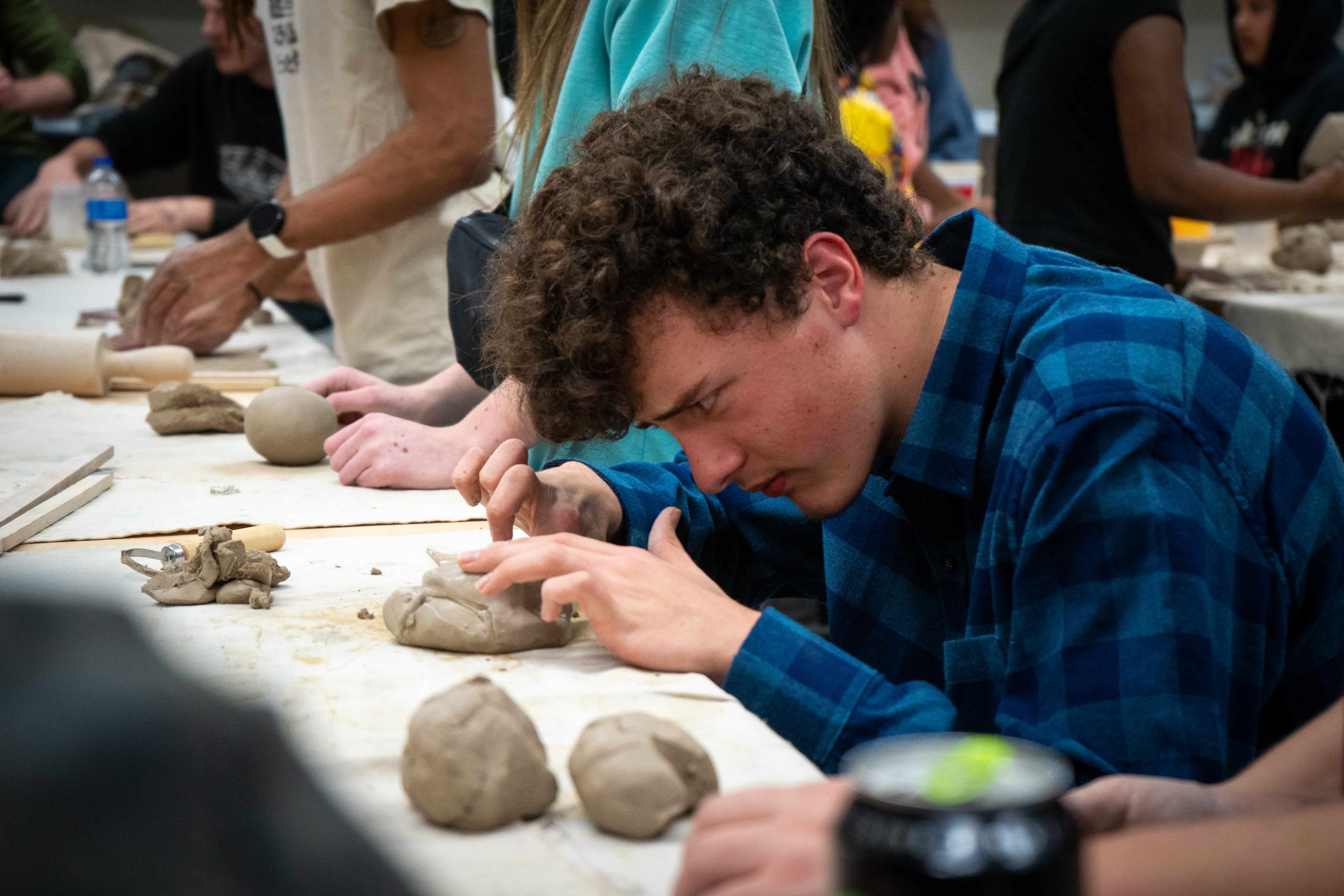 Young male in blue flannel using his hands to mold a lump of clay on the table in front of him.