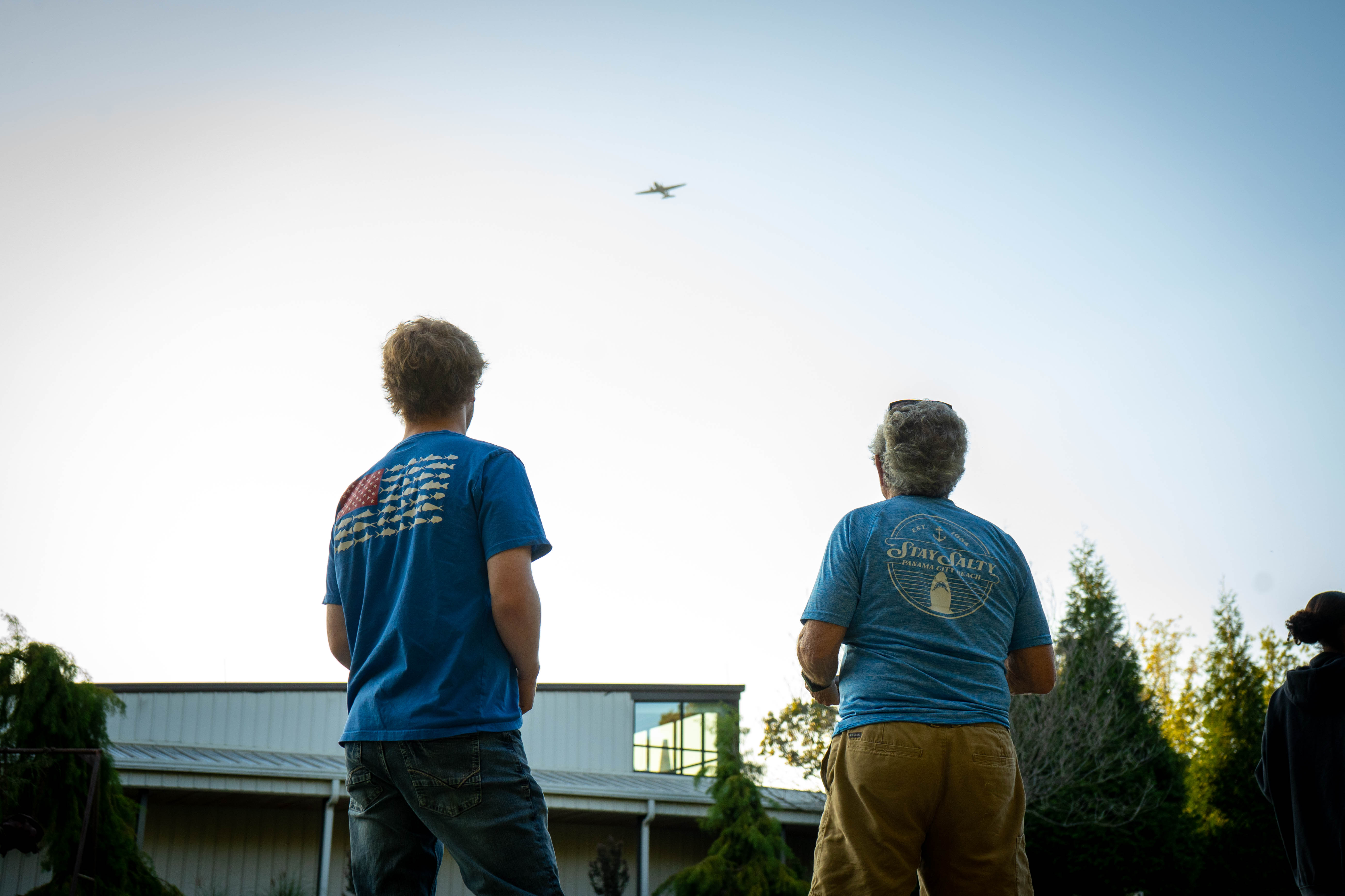 Two people standing together looking up at a plane flying over.
