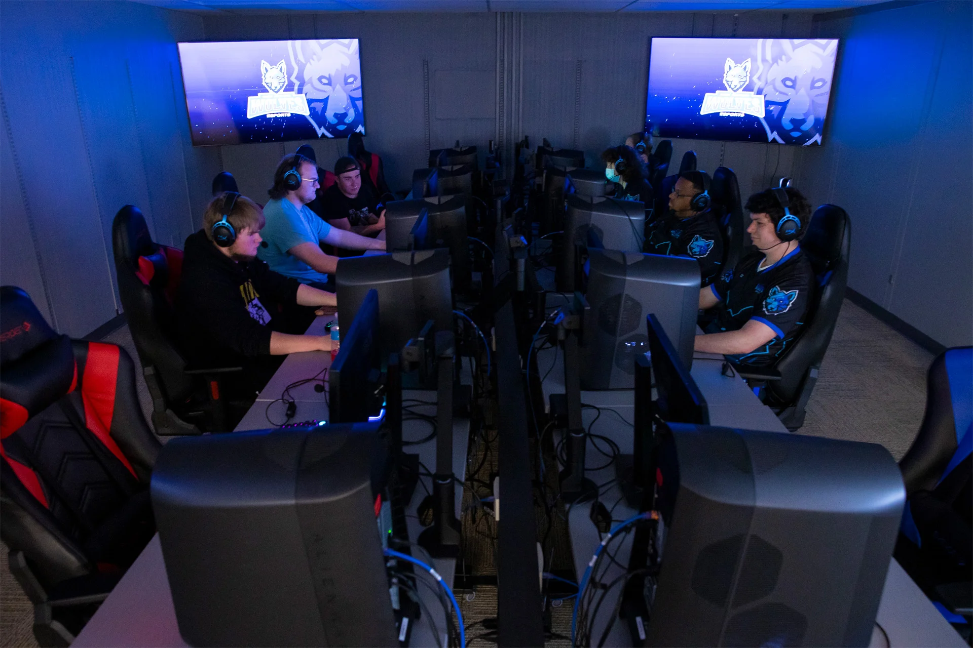 Esports team gathered around computers at a large table with players facing each other at opposite ends of the table.