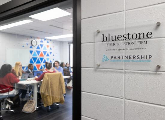 bluestone Public Relations Firm sign in the foreground, looking through the window into the bluestone space at students sitting at tables working together