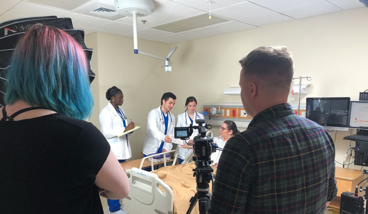 Two individuals filming nursing students in discussion with a patient.