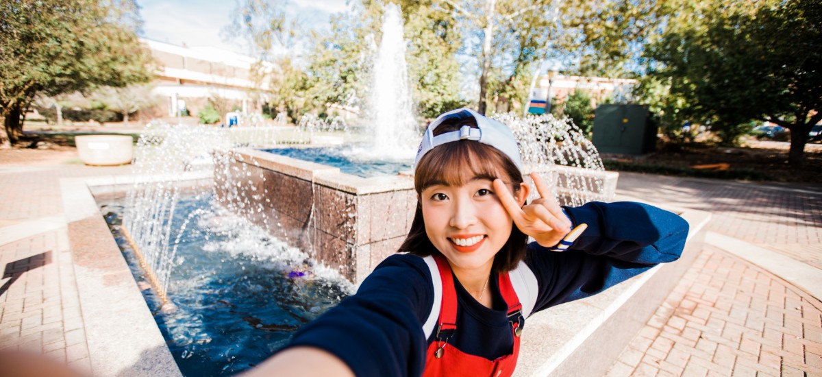 Female student showing the wolf sign in front of the campus fountain.