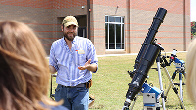 A man in a cap and beard wanders outdoors near some small telescopes