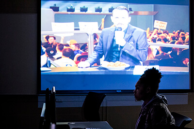 A female student sits in front of a projector screen showing a gaming commentator