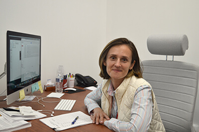 Dr. Stanescu at her office desk with a keyboard, monitior, and sundry papers and other objects.